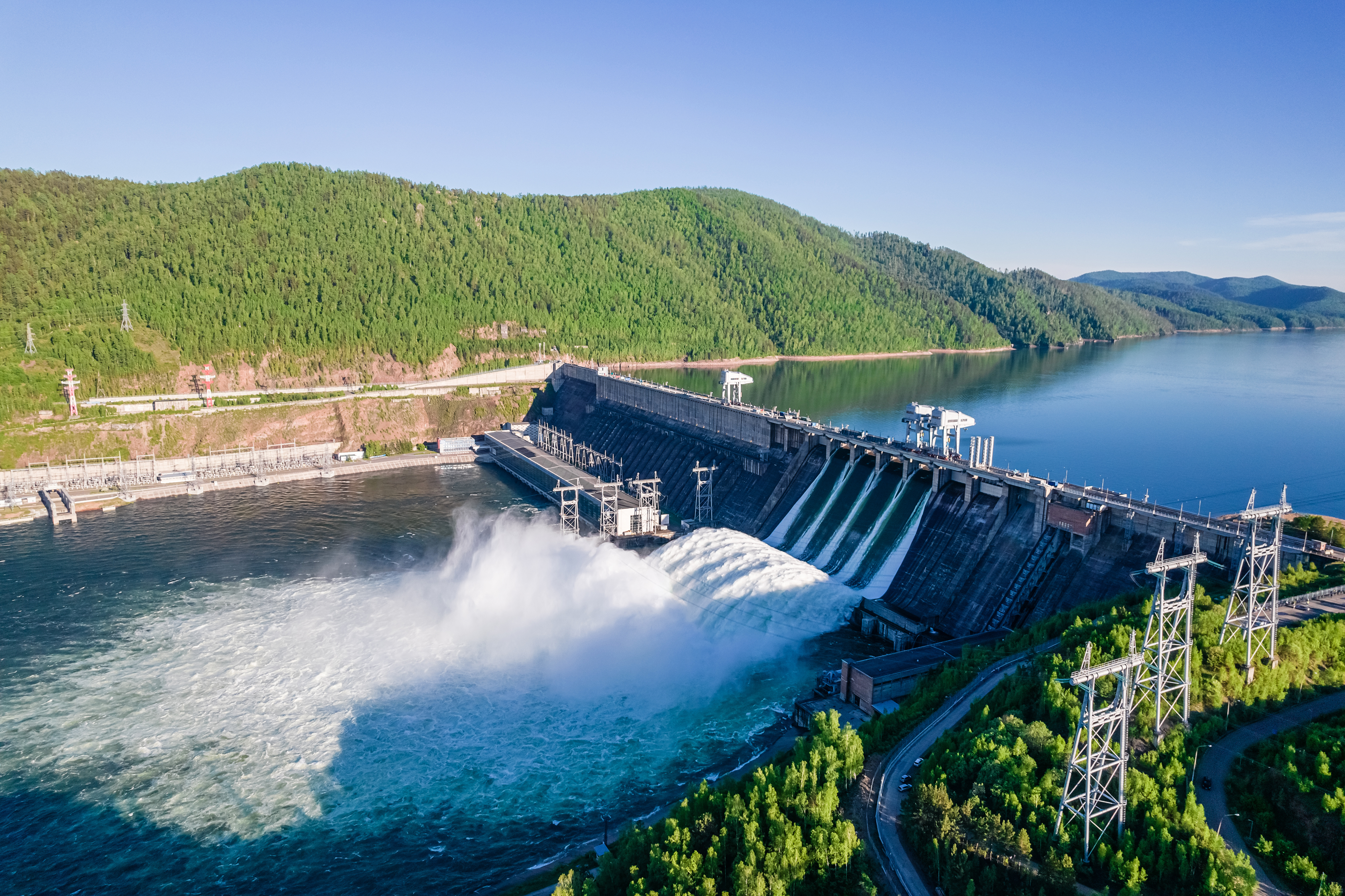 Concrete hydroelectric dam releasing water, with electrical substation and transmission towers visible on the right